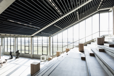 Blick in eine Halle mit stufenförmiger Schautribüne und schräg verlaufender Decke mit Metallbaffeln