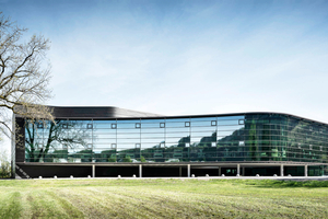  Glatt, grünlich und glänzend liegt der spiegelnde Erweiterungsbau des Firmen-Campus von Graf Elektronik in der Landschaft und lässt diese durch das offene Erdgeschoss fließen 