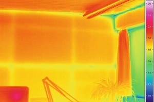  IR-Innenaufnahme (Infrarot-Thermographieaufnahme): Auch der Rollladenkasten über dem Fenster rechts im Bild wurde mit VIP gedämmt. Deutlich sind die Wärmebrücken im Bereich der Anschlussstellen vom Rollladenkasten sowie im Anschlussbereich der Wand mit der Decke zu erkennen. Die deutlich sichtbare waagerecht durch das Bild verlaufende Wärmebrücke in der Wandfläche kommt durch eine Holzlatte zustande, die hier als Befestigungsmöglichkeit mit in die Konstruktion integriert wurde, um beispielsweise Regalkonstruktionen mit der Wand verbinden zu können. Die vertikalen leicht sichtbaren „Wärmebrücken“ sind durch die Stoßkanten der kaschierten vorgefertigten Wandelemente verursacht worden. Um das Beschädigungsrisiko für die VIP im Bereich der Kanten zu reduzieren, wurde die Kaschierung jeweils leicht überstehen gelassen und der Zwischenraum der vorgefertigten Wandelemente mit einem Fugenband ausgeführt. Im oberen Bereich, etwa 40 cm unterhalb der Decke, sieht man schwach ausgeprägte waagerechte Wärmebrücken. Diese ergeben sich allein aus dem Stoß zwischen zwei VIP innerhalb des vorgefertigten Wandelementes 