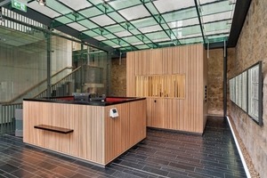  In das Foyer, dessen Hülle und Unterkonstruktion aus Glas bestehen, sind freistehende Betonkuben gestellt, die mit Holz verkleidet sind 