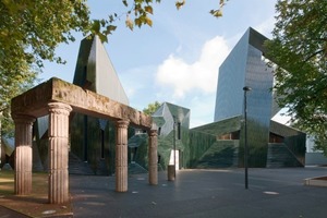  Blaugrün schimmert die Keramikfassade durch die Bäume der Mainzer Neustadt. Im Vordergrund erinnern Mauerreste an die alte, von den Nationalsozialisten zerstörte Synagoge 